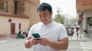 Young chinese man smiling confident using smartphone at street