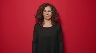 Orta yaşlı bir kadın gülümsüyor. Kollarını kavuşturup kırmızı arka plan üzerinde hareketler yapıyor.