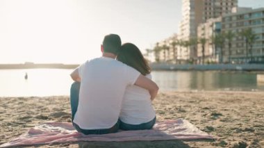 Kadın ve erkek kumsalda havlunun üzerinde oturup birbirlerine sarılıyorlar.