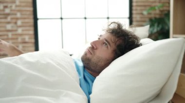 Young hispanic man lying on bed covering ears with pillow for noise at bedroom