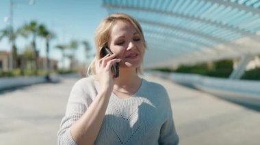 Genç sarışın kadın gülümsüyor. Parktaki akıllı telefondan konuşuyor.