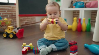 Adorable hispanic baby playing with construction blocks sitting on floor at kindergarten