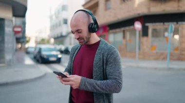 Genç kel adam gülerek sokakta müzik dinliyor.