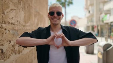 Genç beyaz adam gülümsüyor. Kendine güveniyor. Elleriyle kalp hareketi yapıyor.