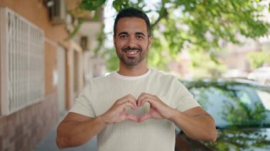 Young hispanic man smiling confident doing heart gesture with hands at street