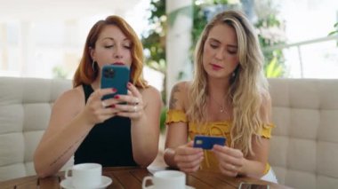 Two women using smartphone and credit card sitting on table at home terrace