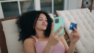 Akıllı telefon ve kredi kartı kullanan genç İspanyol kadın terasta güverte sandalyesinde yatıyor.