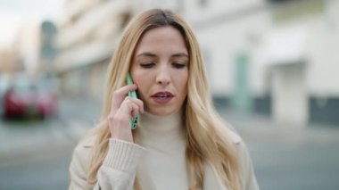 Ciddi bir suratı olan genç sarışın kadın caddede akıllı telefondan konuşuyor.