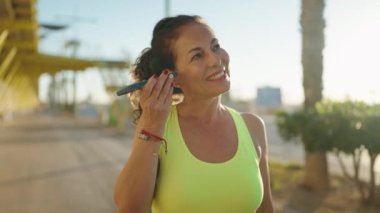 Orta yaşlı kadın spor giyer ve caddedeki akıllı telefondan sesli mesaj dinler.