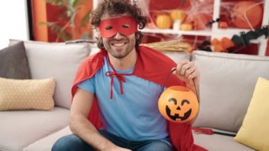 Young hispanic man wearing superhero costume holding halloween pumpkin basket at home