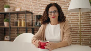 Orta yaşlı bir kadın evde kahve içerken kendinden emin gülümsüyor.