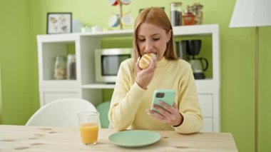 Young blonde woman using smartphone having breakfast at home