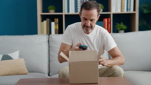 家でスマートフォンの段ボールの箱をアンパックを使用して中年の男 — ストック動画
