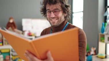 Young hispanic man preschool teacher reading book standing at kindergarten