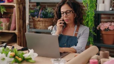 Middle age woman florist talking on smartphone using laptop at flower shop