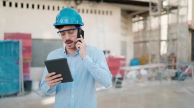 Young hispanic man architect talking on the smartphone using touchpad at street