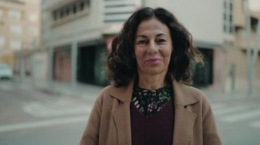 Orta yaşlı İspanyol kadın gülümsüyor. Kollarını kavuşturup caddede duruyor.