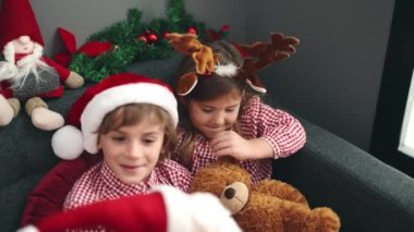 Erkek ve kız kardeş oyuncak ayı ve Noel Baba oyuncağıyla oynuyorlar. Evdeki Noel süslemesinde koltukta oturuyorlar.