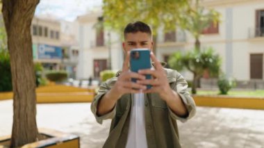 Genç İspanyol adam gülümsüyor. Parktaki akıllı telefonun yanında özçekim yapıyor.