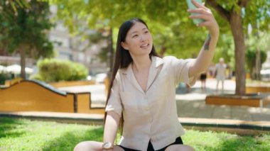 Parktaki çimlerin üzerinde oturan Çinli kadın akıllı telefonun yanında selfie çekiyor.