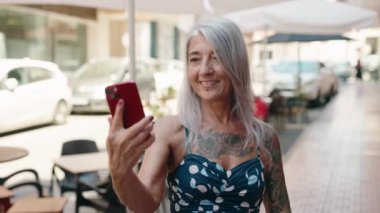 Middle age grey-haired woman smiling confident making selfie by the smartphone at street