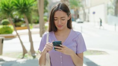 Genç, güzel, İspanyol bir kadın sokakta akıllı telefon kullanarak gülümsüyor.