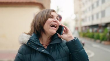 Orta yaşlı bir kadın gülümsüyor. Sokakta akıllı telefondan konuşuyor.