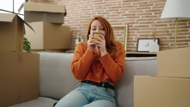 年轻的红头发女人坐在新家的沙发上喝咖啡 — 图库视频影像