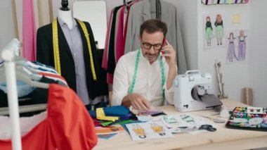 Orta yaşlı bir terzi terzide elinde kıyafetlerle akıllı telefondan konuşuyor.