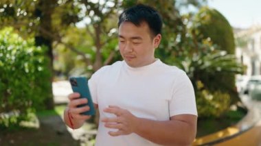 Genç Çinli adam gülümsüyor. Parktaki akıllı telefonun yanında özçekim yapıyor.