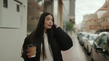 Young hispanic woman talking on the smartphone drinking coffee at street