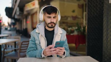 Young hispanic man listening to music sitting on table at coffee shop terrace