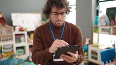 Young hispanic man preschool teacher writing on touchpad standing at kindergarten