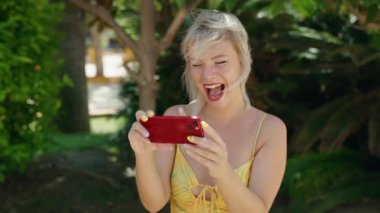 Genç sarışın kadın gülümsüyor. Parkta akıllı telefondan videolar izliyor.