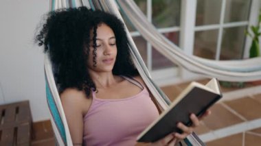 Young hispanic woman reading book lying on hammock at home terrace