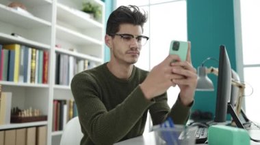 Young hispanic man student using smartphone sitting on table at library university