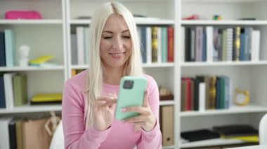 Young blonde woman student using smartphone sitting on table at library university