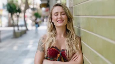 Genç sarışın kadın gülümsüyor. Kollarını kavuşturup caddede duruyor.