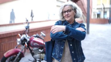 Orta yaşlı, gri saçlı, kolları çapraz caddede gülümseyen bir kadın.