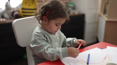 Anaokulunda masaya oturmuş kağıda resim çizen sevimli İspanyol kız öğrenci.