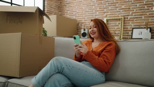 在新家用智能手机坐在沙发上的红头发年轻女人 — 图库视频影像