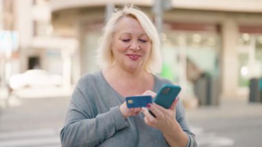 Middle age blonde woman using smartphone and credit card at street