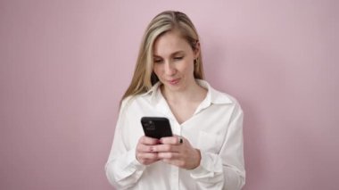 Young blonde woman using smartphone standing over isolated pink background