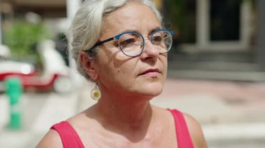 Orta yaşlı, kır saçlı bir kadın sokakta ciddi bir ifadeyle yan tarafa bakıyor.