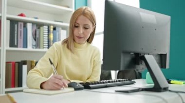 Young blonde woman student using computer writing on notebook at university classroom