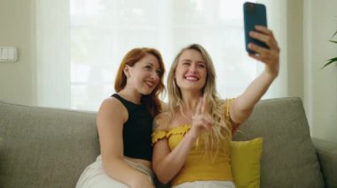 Two women making selfie by smartphone sitting on sofa at home