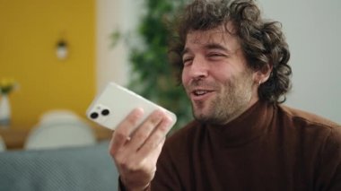 Young hispanic man talking on the smartphone sitting on sofa at home