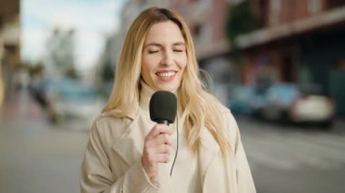 Young blonde woman journalist speaking using microphone at street