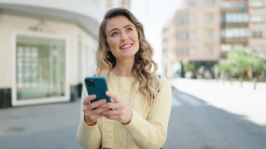 Genç sarışın turist sokakta akıllı telefon kullanarak gülümsüyor.