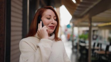 Genç kızıl saçlı, gülümseyen, kendine güvenen, caddedeki akıllı telefondan konuşan kadın.
